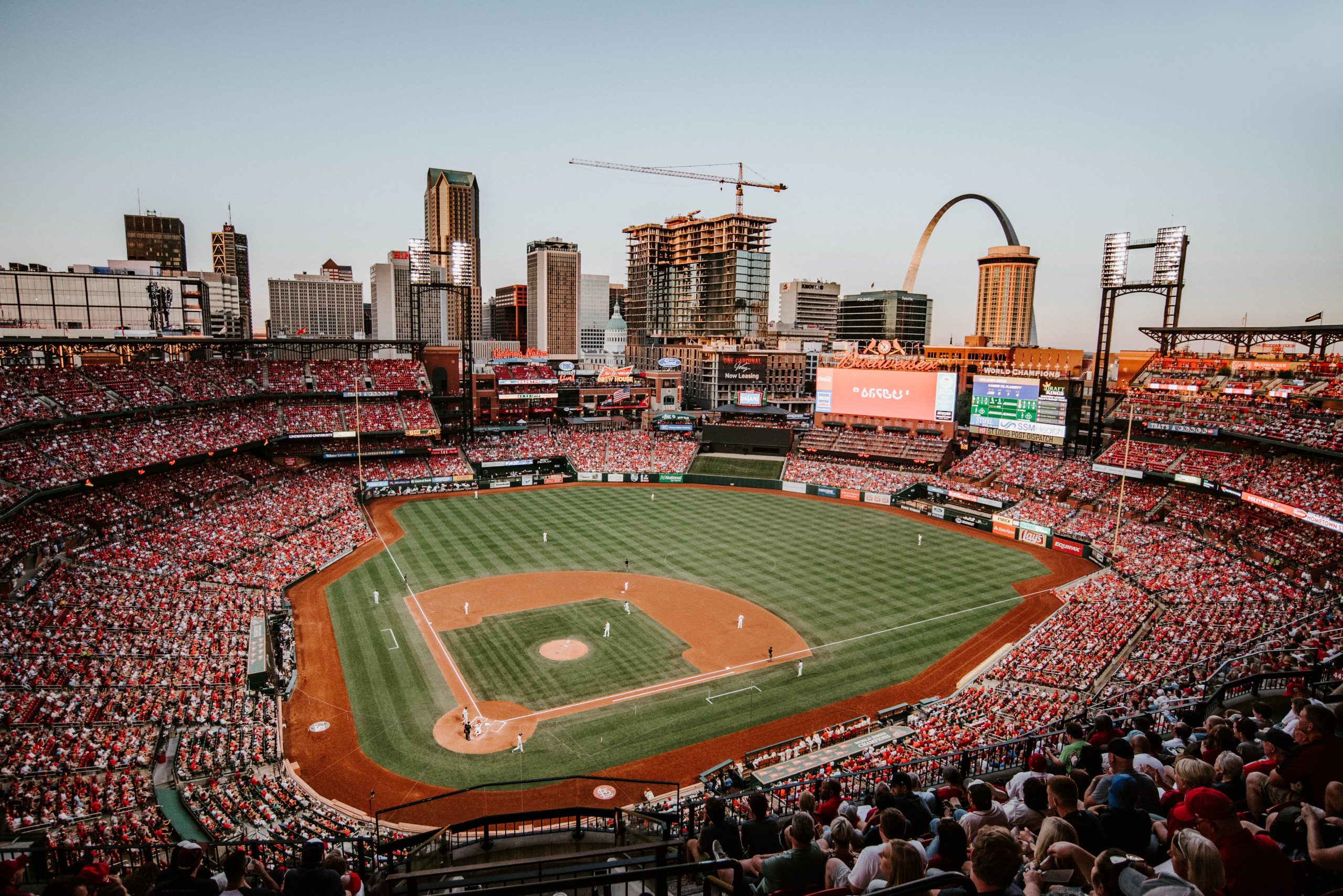Photograph of Busch Stadium in St. Louis, filled with fans. A Cardinals game is underway, and the St. Louis arch is visible in the background.