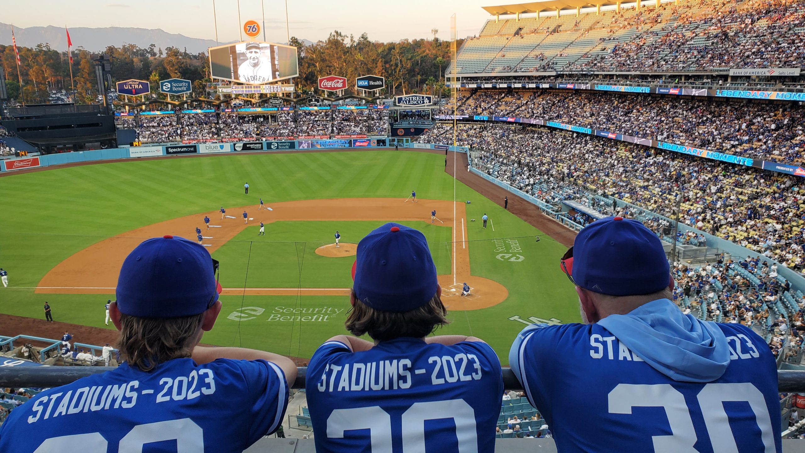 Photograph of the backs of Ryan, Heather and Brad at Dodger Stadium, wearing jerseys that say "Stadiums - 2023: 30" and looking down over the baseball diamond.